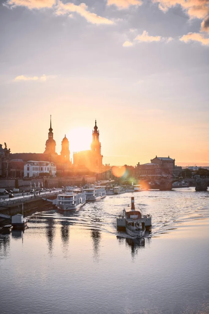 Sonnenuntergang über Stadtfluss mit historischen Gebäuden und vorbeifahrenden Booten im Vordergrund.
